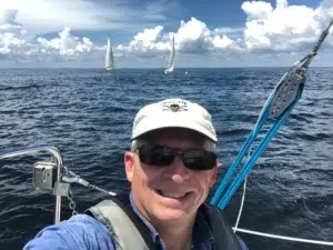 Man wearing a cap and sunglasses on a sailboat, smiling with the open sea and distant sails behind him, blue sky above.